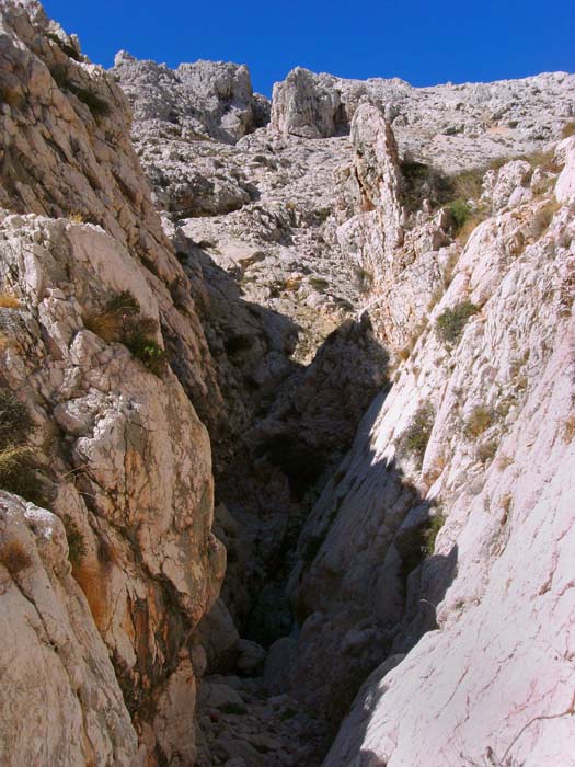 wo der Weg rechts aus dem Canyon emporsteigt, kann man durch ein leicht zu bewegendes Metallsperrgitter weiter vordringen; oben schon deutlich sichtbar die „Gipfelschlucht“; nach dieser Engstelle mit gut mannshoher Kletterpassage biegt der Spalt 90° nach rechts und erweitert sich etwas (Steinmann); dort verlässt man den Schluchtgrund nach links
